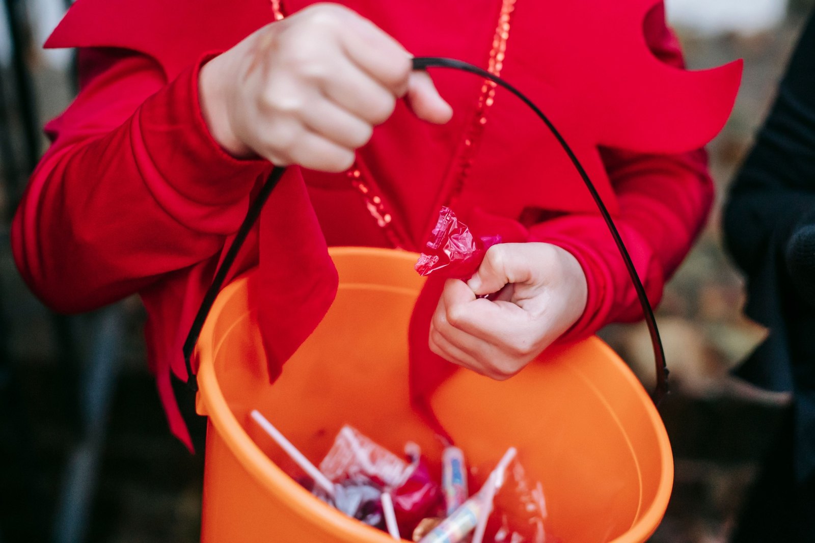 A child in a red costume reaching into an orange Halloween candy bucket